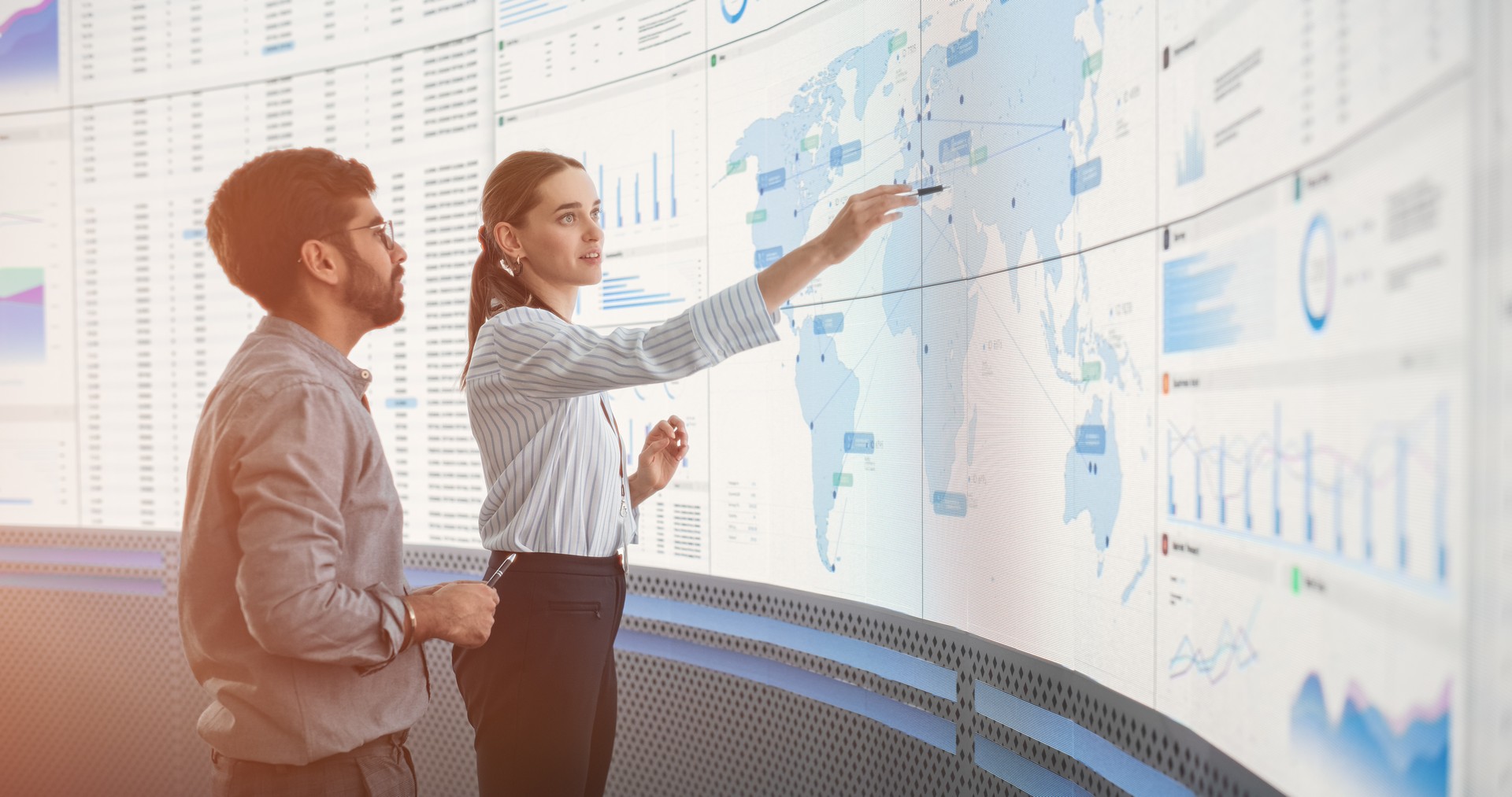 Indian Male Data Scientist And Caucasian Female Business Manager Talking In Front Of Big Digital Screen With Graphs, Charts And Map In Monitoring Office. Diverse Colleagues Discussing Objectives.